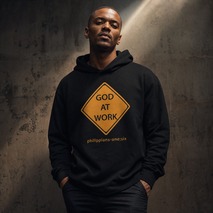 Man wearing a black t-shirt hoodie with a caution sign design with 'God At Work' text standing against a gray wall.