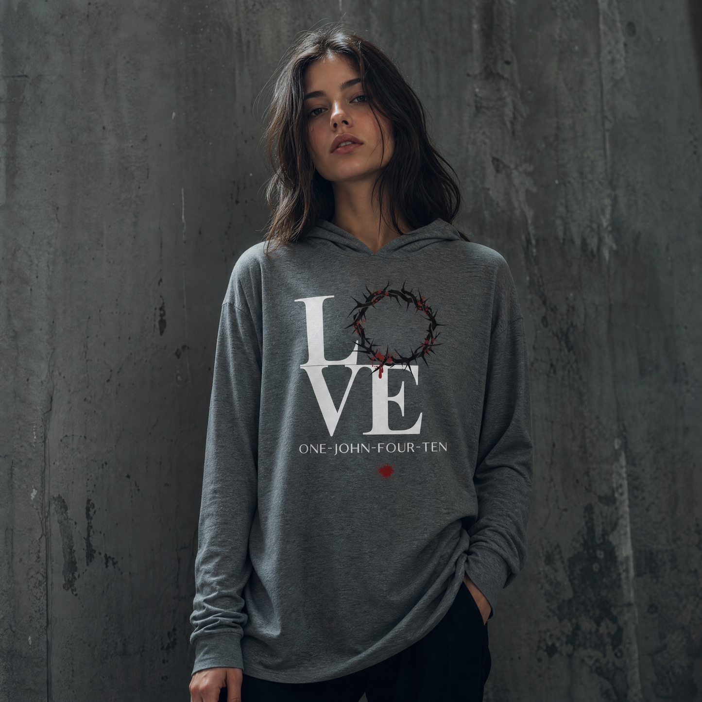 Model is wearing a gray long sleeve t-shirt hoodie with text 'LOVE' and a graphic of the crown of thorns in place of the 'O' standing in front of a gray wall.