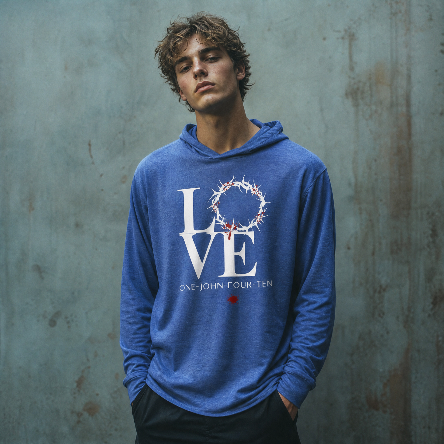 Model is wearing a heather royal blue long sleeve t-shirt hoodie with text 'LOVE' and a graphic of the crown of thorns in place of the 'O' standing in front of a gray wall.
