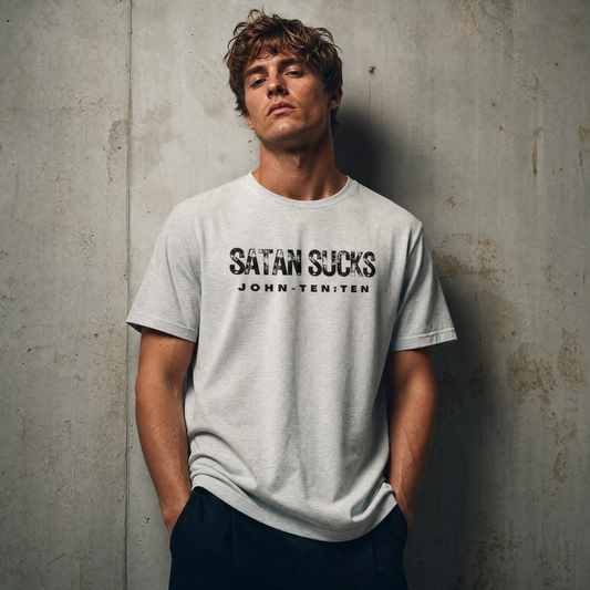 A man wearing a light gray t-shirt with the text 'SATAN SUCKS' printed in black across the chest, standing against a textured concrete wall.