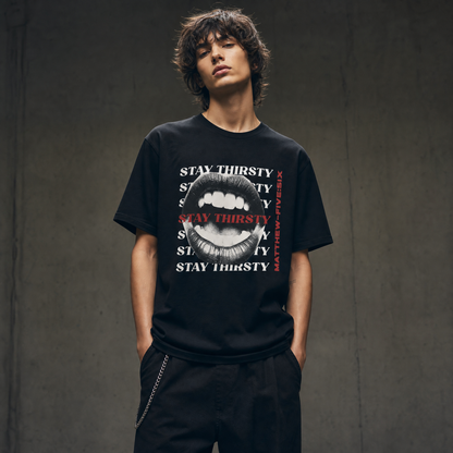 Model wearing a black t-shirt with text 'Thirsty' and an open mouth graphic; standing against a concrete wall