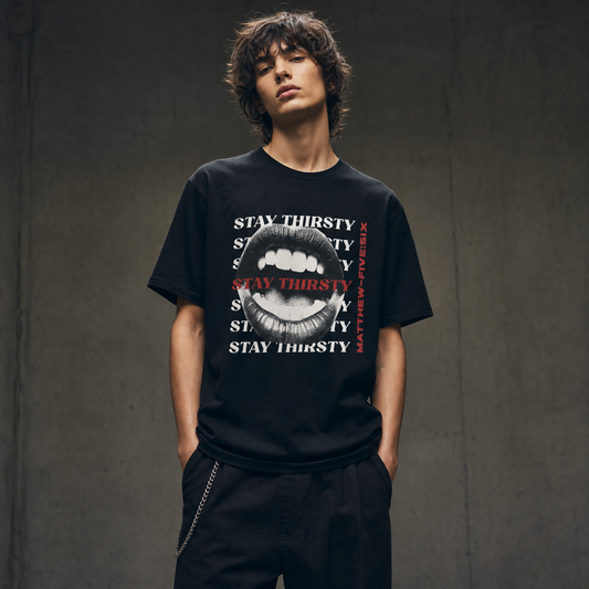 Model wearing a black t-shirt with text 'Thirsty' and an open mouth graphic; standing against a concrete wall