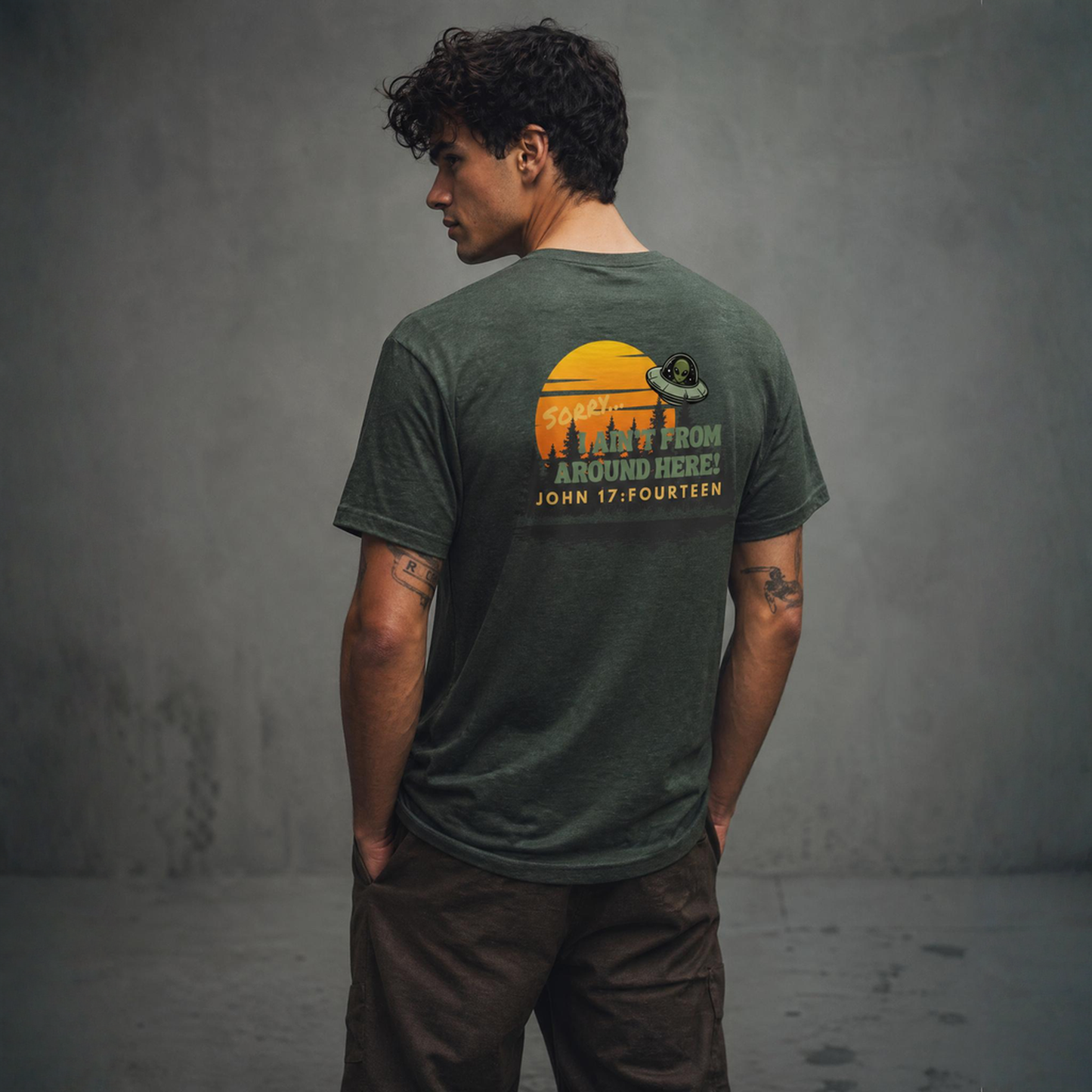 Model wearing a heather forest green t-shirt with graphic and text 'Sorry, I Ain't From Around Here' standing next to a concrete wall