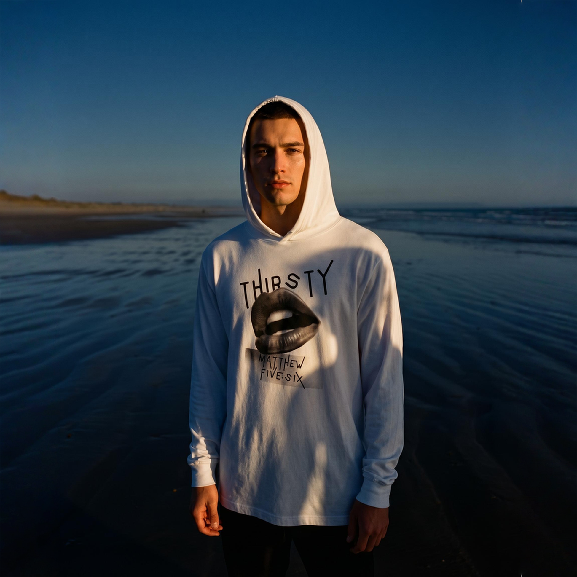Person standing on a beach wearing a white t-shirt hoodie with text Thirsty 