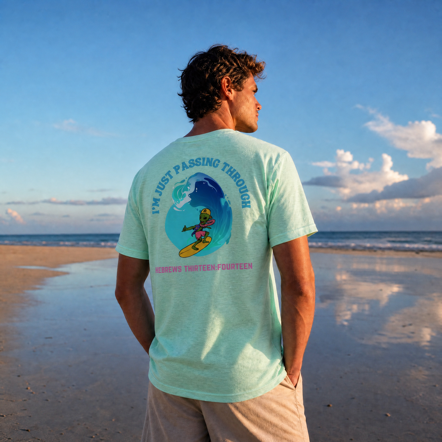 Man standing on the beach wearing a heather mint green t-shirt with a graphic  of a surfing alien with the words I'm Just Passing Through 